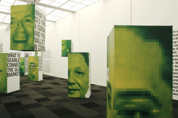 Pedestals, some on the floor, some hanging from the ceiling, display large green portraits of various people in a white gallery space with dark gray carpet