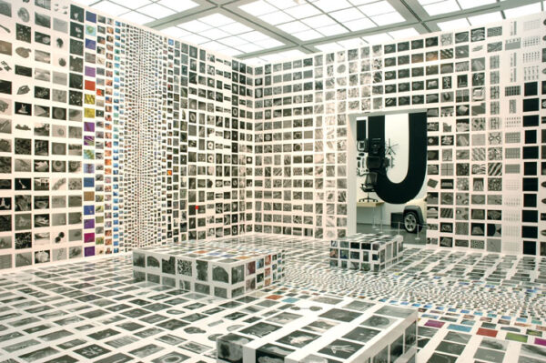 Hundreds of pictures cover the walls, floor, and furniture of a gallery space