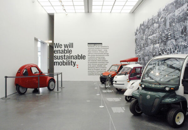 Various mini cars in a white gallery space with black text on the far white wall. The legible text reads: We will enable sustainable mobility.