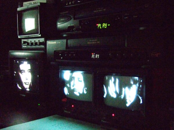 Four TV screens in a dark room, three on the bottom displaying various black and white images of a woman's face; the fourth sits onto of the first tv, the screen is dark