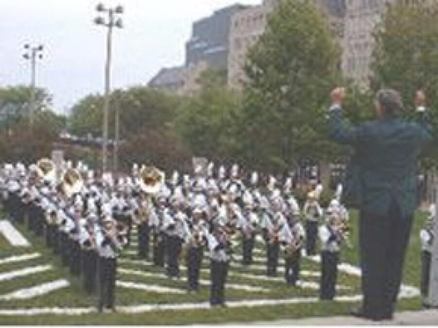 A conductor faces a marching band lined up in four rows in the MCA's backyard. The academy records logo appears beneath the image