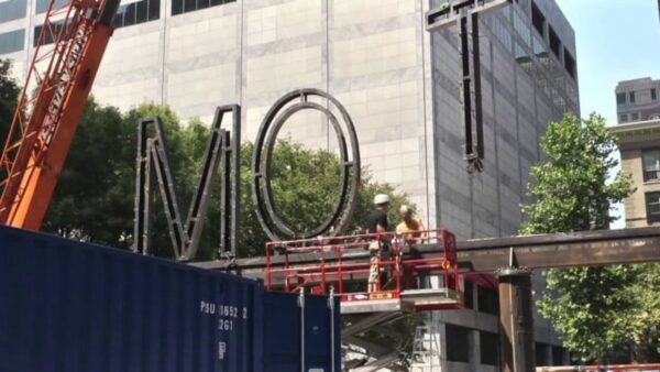 A crane lifts a large letter T onto a metal beam which already has the letters M and O on it