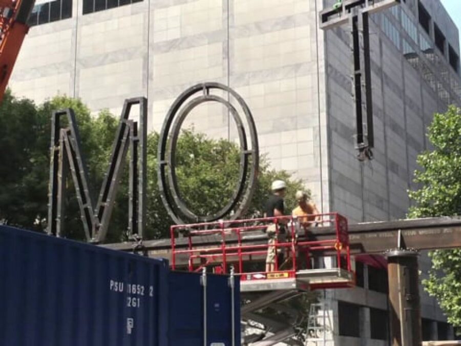 A crane lifts a large letter T onto a metal beam which already has the letters M and O on it