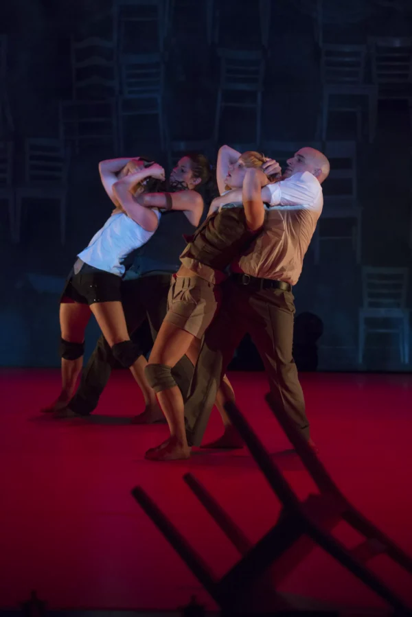 Performance still of two people putting two other people in a chokehold on a red-lit stage with a background of chairs. In the foreground a chair has been tipped over.