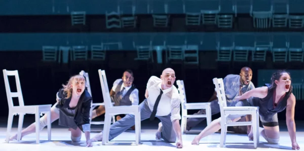 Performance still of five performers lunging next to white chairs with aggressive looks on their faces.