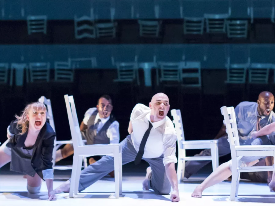 Performance still of five performers lunging next to white chairs with aggressive looks on their faces.