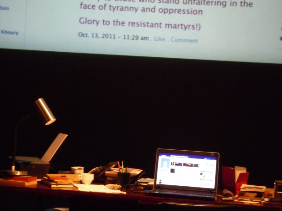 A desk with a lamp and laptop computer on a dark stage with a large screen displaying the computer screen at a giant scale