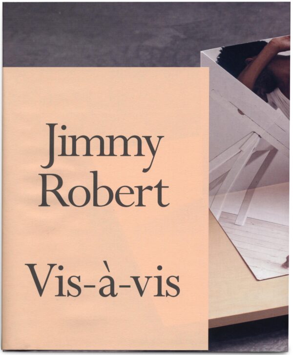 A peach piece of paper with black font reading Jimmy Robert, Vis-a-vis covers an image of paper folded on a wooden base