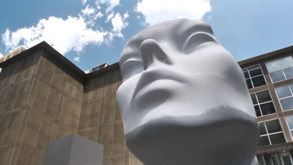 The front of a giant mannequin head as viewed from below that is painted grey from the chin down with a white face