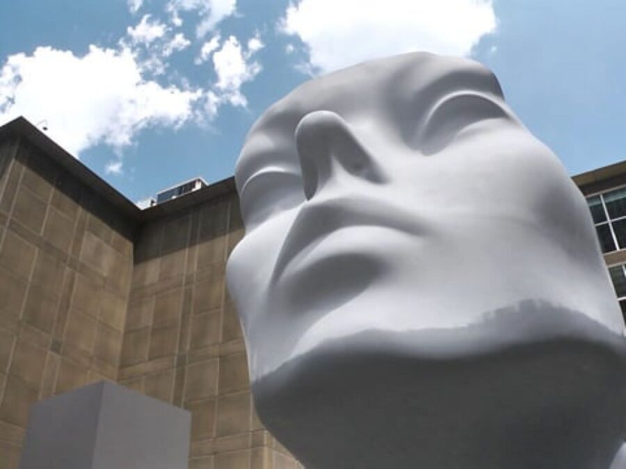 The front of a giant mannequin head as viewed from below that is painted grey from the chin down with a white face