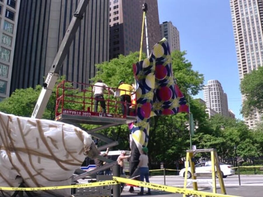 A crane and an industrial lift carrying two people work on positioning a large, colorful object in an area near skyscrapers