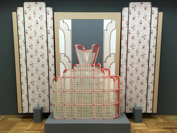 The armature of a hoop dress on a soft blue grey pedestal against a backdrop of faux art deco columns in a floral pattern