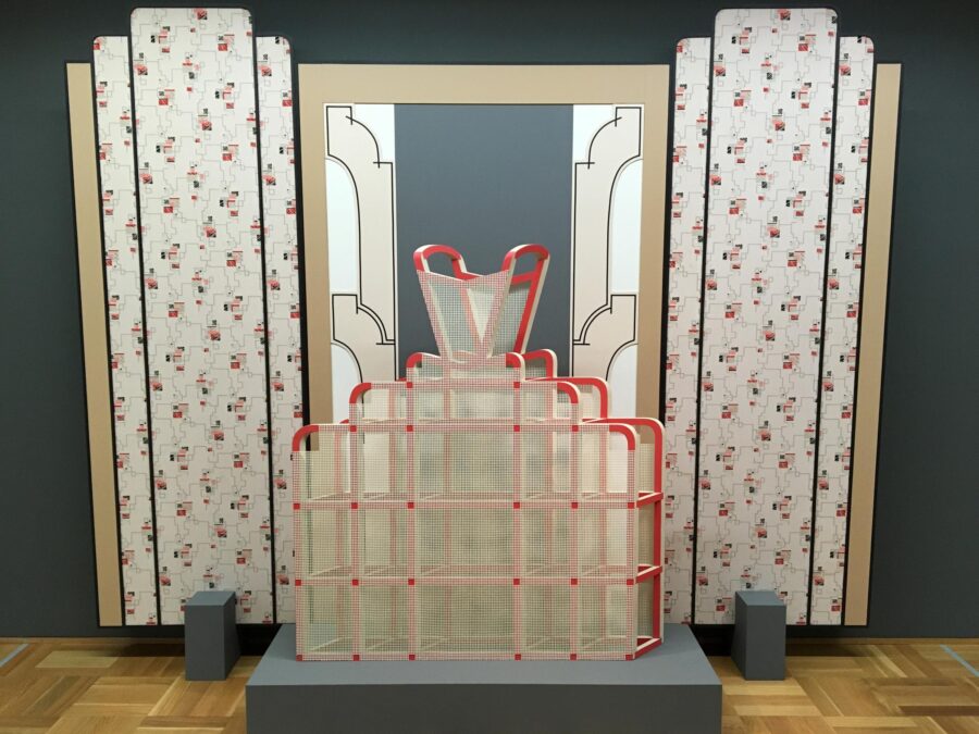 The armature of a hoop dress on a soft blue grey pedestal against a backdrop of faux art deco columns in a floral pattern