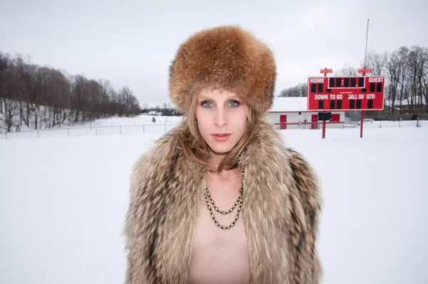 A person in a fur hat and coat stands with their coat open, revealing a bare chest, in a snow-covered field with a scoreboard in the background