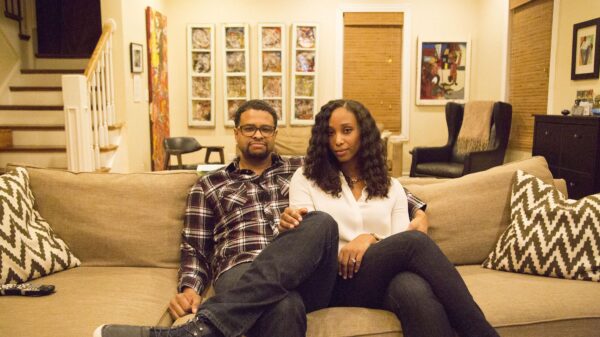 A man and a woman sit close to each other on a couch in a homey room decorated with a lot of colorful art