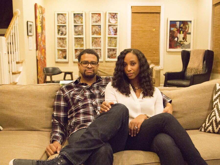 A man and a woman sit close to each other on a couch in a homey room decorated with a lot of colorful art