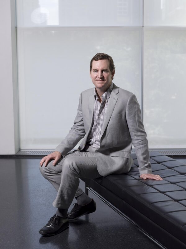 A male presenting person with light-toned skin in a grey suit sitting on a black bench