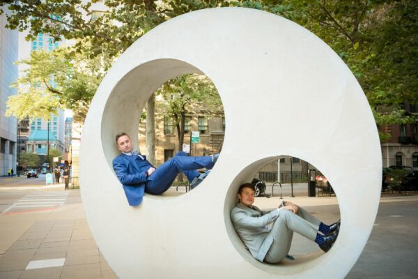 Two male-presenting people in suits pose in holes cut into a large concrete circle.