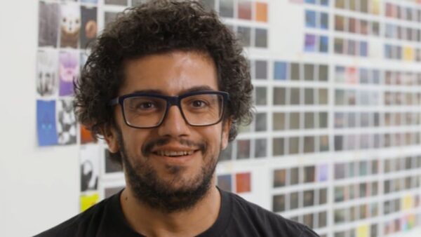 A man with light brown skin, dark curly hair, and dark glasses smiles in front of a wall filled with small square photos