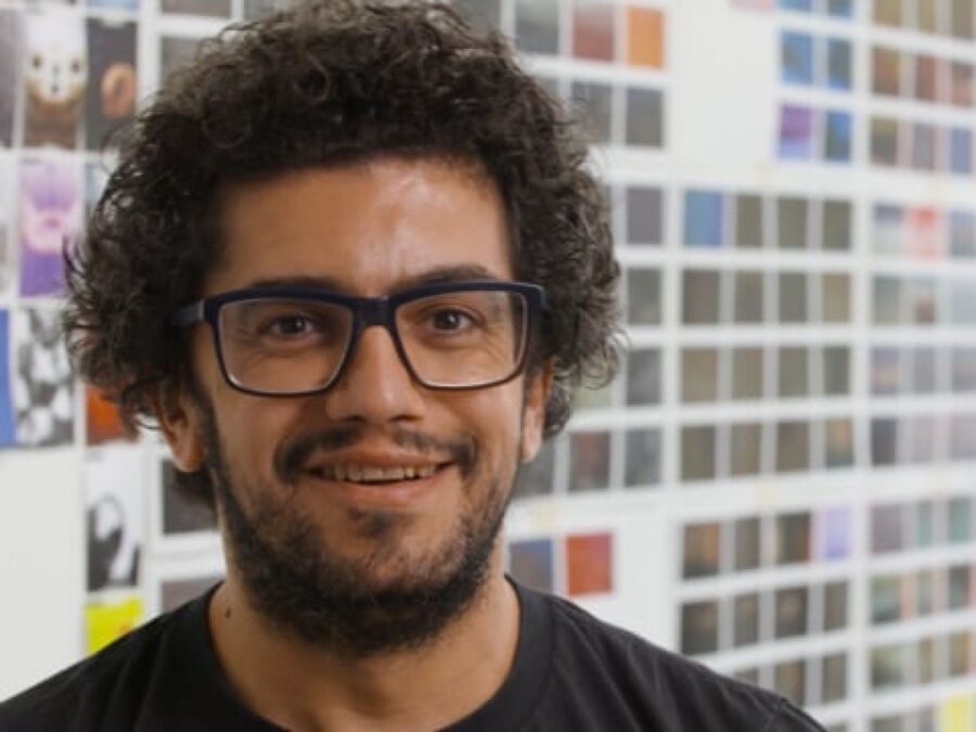 A man with light brown skin, dark curly hair, and dark glasses smiles in front of a wall filled with small square photos