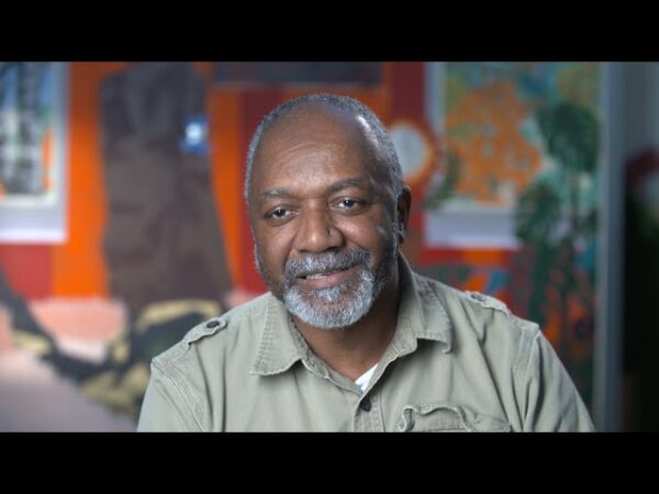 A Black man with a gray beard smiles in front of a large artwork