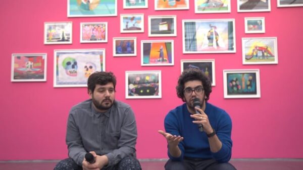 Two people sit in front of a magenta wall filled with various small photographs in white frames. The man on the right holds the microphone up to their moth and gestures with their other hand.