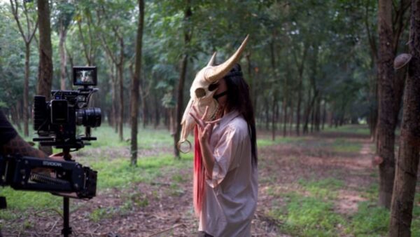 A person in a horned-animal skull mask poses in front of a camera in a forest