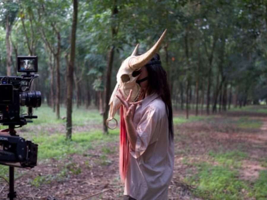A person in a horned-animal skull mask poses in front of a camera in a forest
