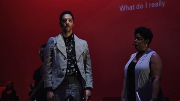 Three people in front of a red screen with grey text reading What do I really. The two actors in the foreground are dressed in grey and black outfits. Behind the actor on the left, a third person dressed in black is just visible.