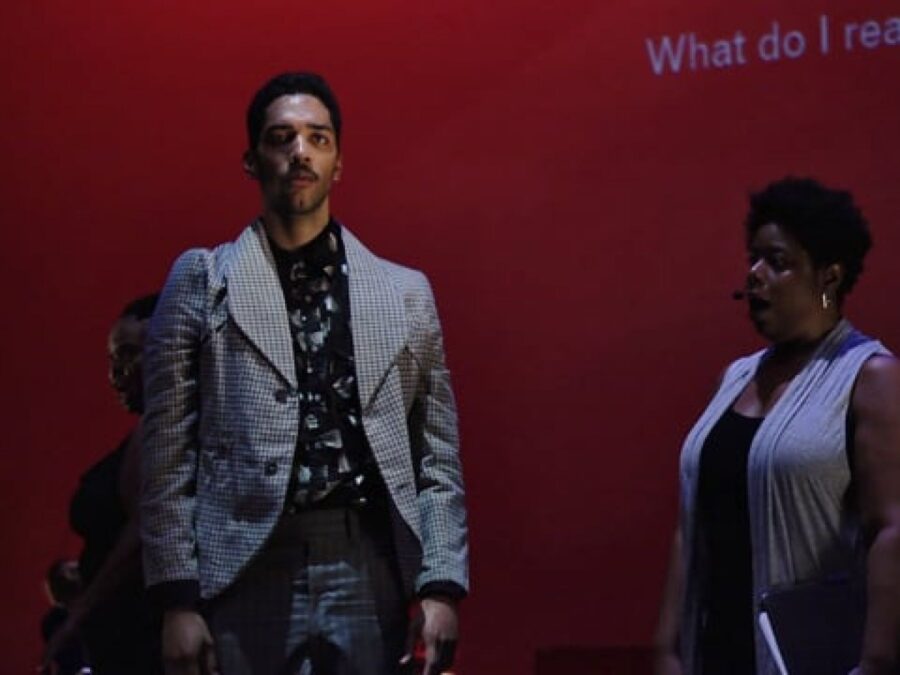 Three people in front of a red screen with grey text reading What do I really. The two actors in the foreground are dressed in grey and black outfits. Behind the actor on the left, a third person dressed in black is just visible.