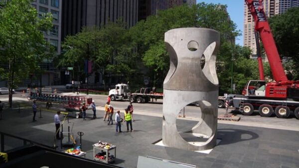 Construction crews near a large concrete pipe with holes in it placed upright on a stone plaza