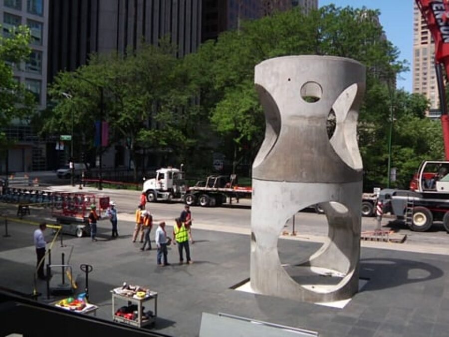 Construction crews near a large concrete pipe with holes in it placed upright on a stone plaza