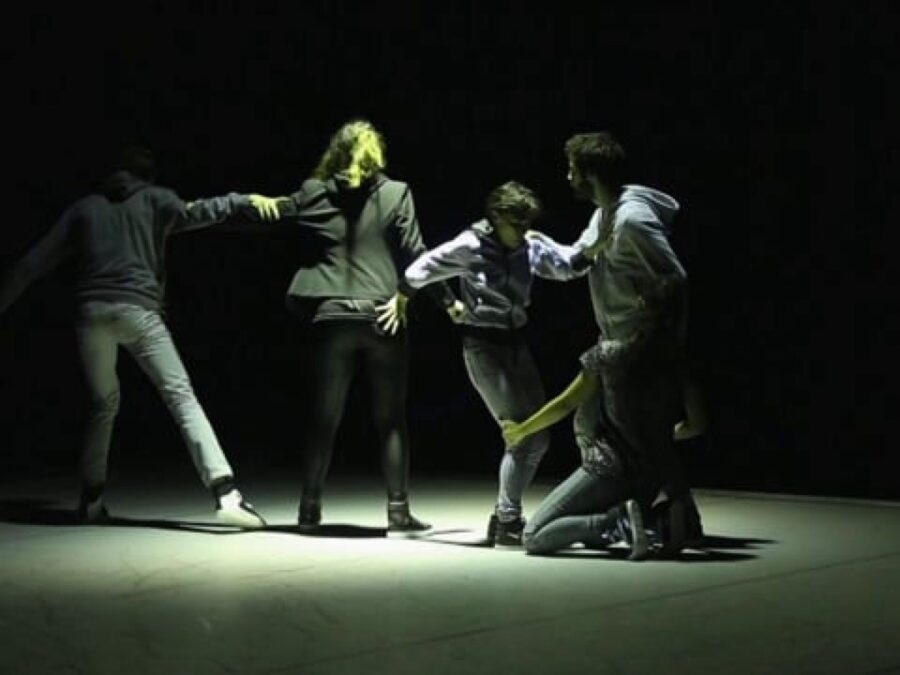 Four people hold onto each other on a dark stage with one spotlight