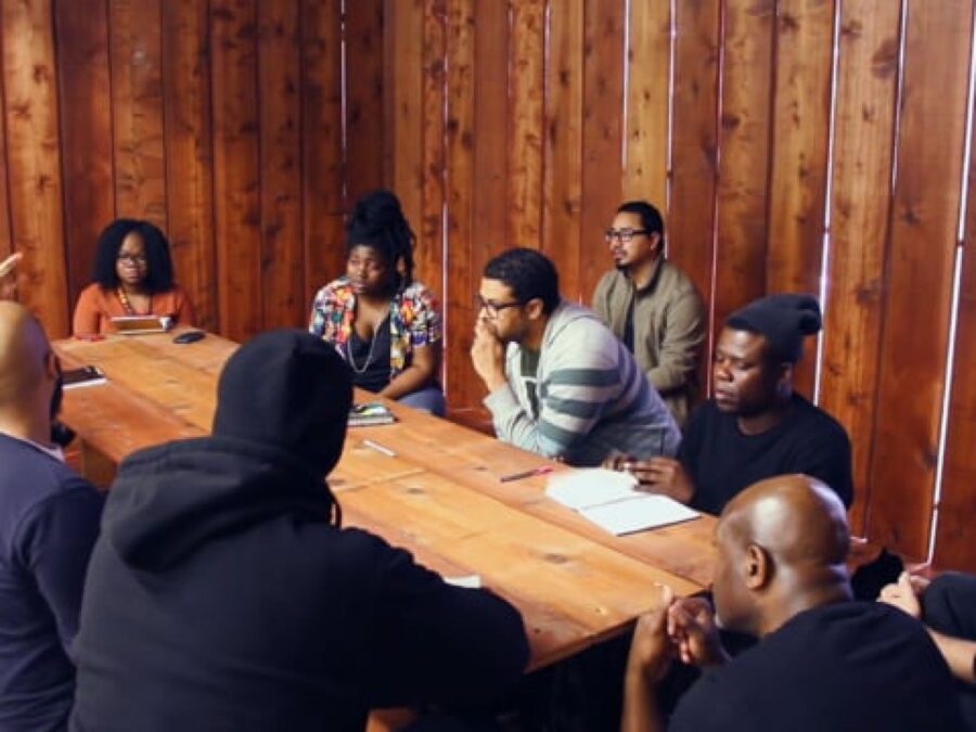 A group of people sit around a wooden table in a wood-paneled room
