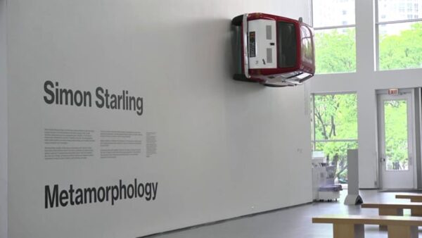 A red and white car hangs on a wall near black text that reads Simon Starling Metamorphology in a large gallery space with doors leading to the outside in the background