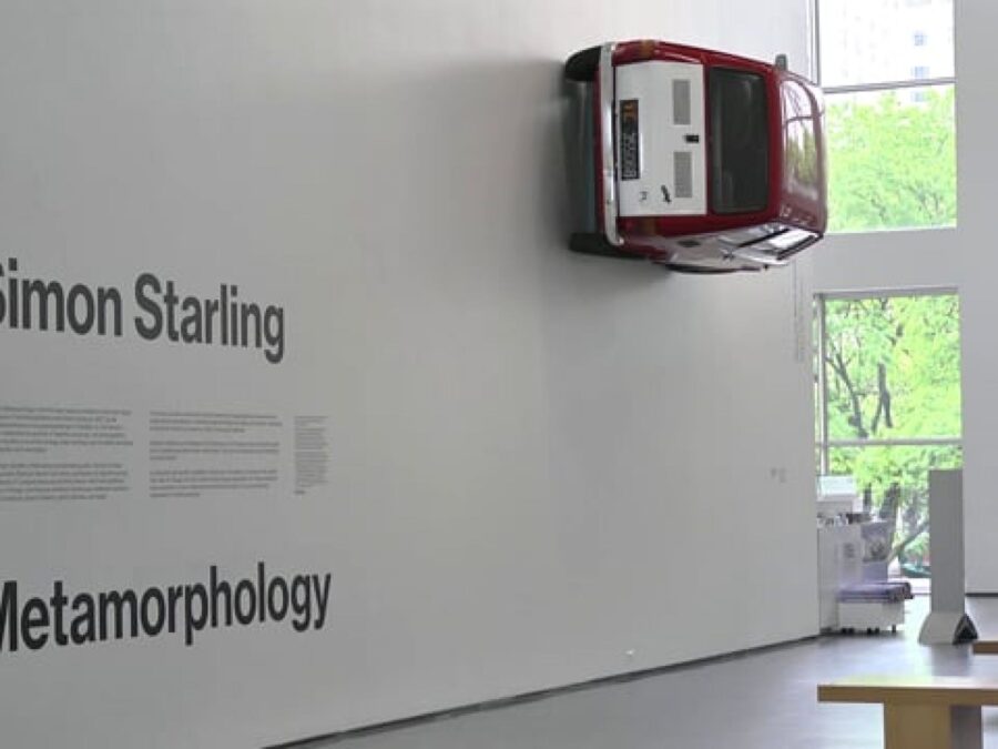 A red and white car hangs on a wall near black text that reads Simon Starling Metamorphology in a large gallery space with doors leading to the outside in the background