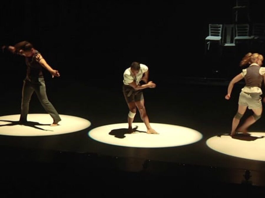 Three dancers in various positions lit only by three spotlights on a dark stage