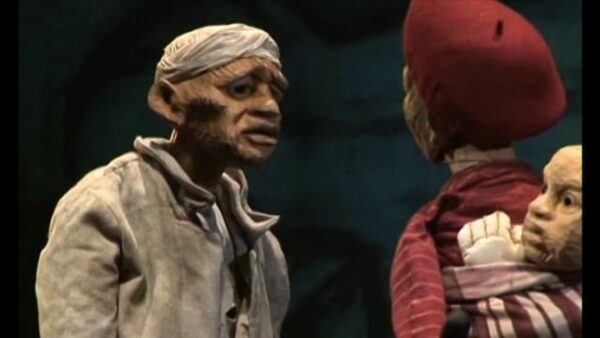 Two puppets, one with a baby on their back, interact on a black set