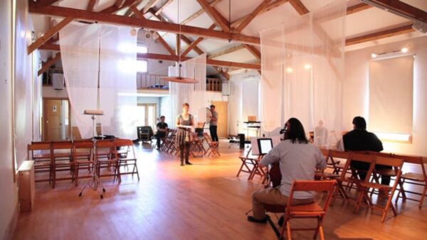 Performance rehearsal in a bright airy space with exposed tresses and sheer, flowy curtains that surround a standing person who holds a book in front of a seated tech crew