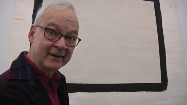 An older gentleman with glasses bends into the camera frame in front of a black square outline