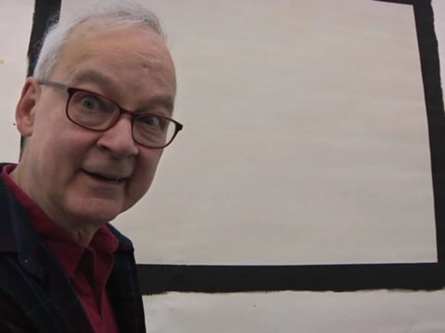 An older gentleman with glasses bends into the camera frame in front of a black square outline