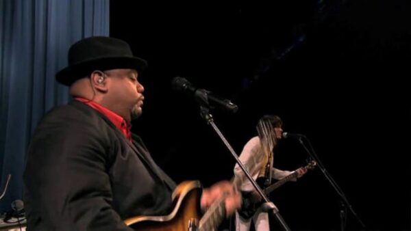 Two electric guitarists on a dark stage standing behind two microphone stands