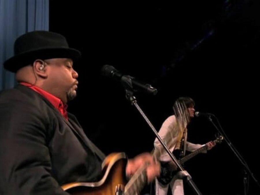 Two electric guitarists on a dark stage standing behind two microphone stands