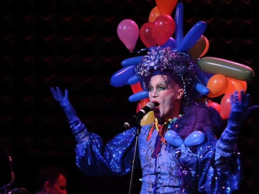 A person in a blue dress with a balloon headdress stands in front of a microphone with their mouth open