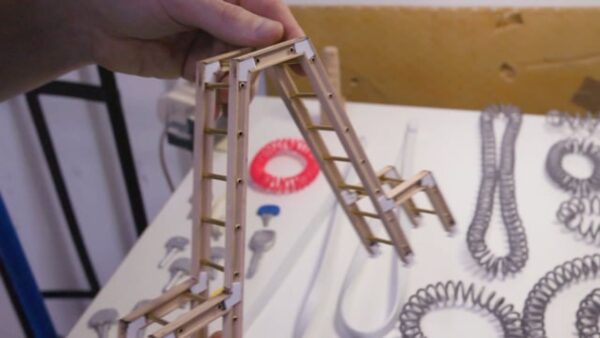 A light-skinned left hand holds a tiny double ladder above a table covered in various sized coils