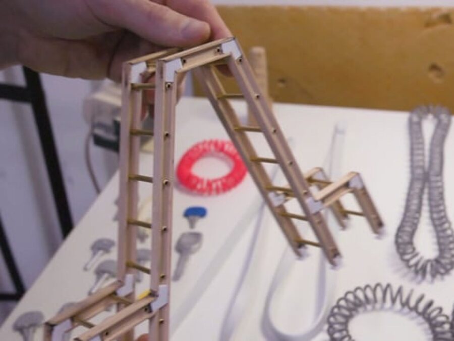 A light-skinned left hand holds a tiny double ladder above a table covered in various sized coils