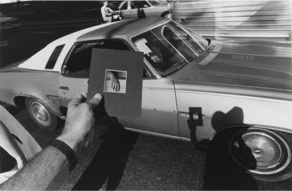A person's left hand holds a picture mat over a car to highlight the passenger's right hand. A shadow of the photographer is cast onto the car
