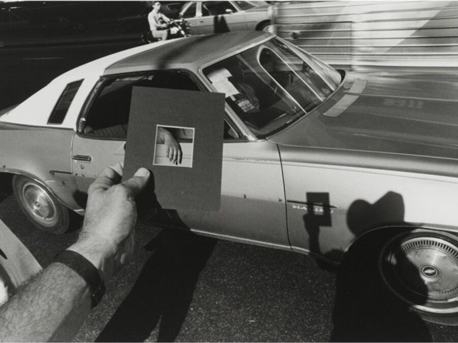 A person's left hand holds a picture mat over a car to highlight the passenger's right hand. A shadow of the photographer is cast onto the car