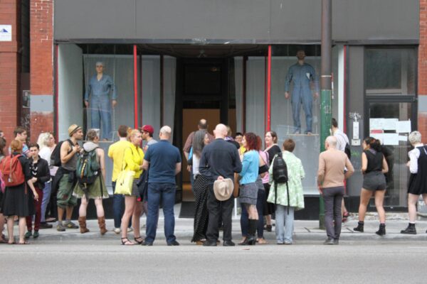 A crowd observes two people dressed in blue jumpsuits standing on makeshift swings in two store windows