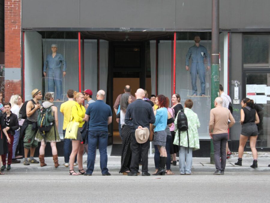 A crowd observes two people dressed in blue jumpsuits standing on makeshift swings in two store windows